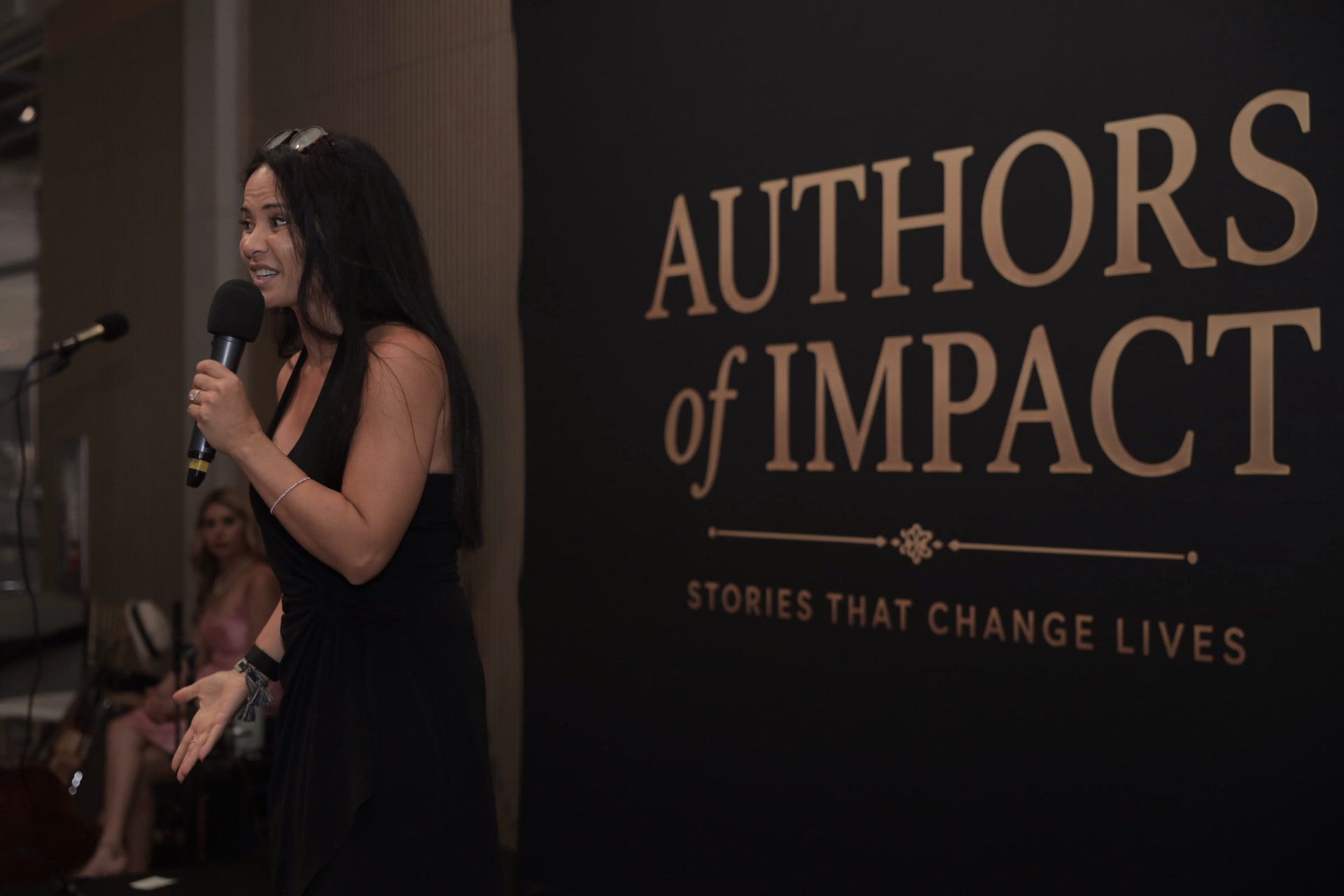 Host speaking on stage in front of the Authors of Impact backdrop